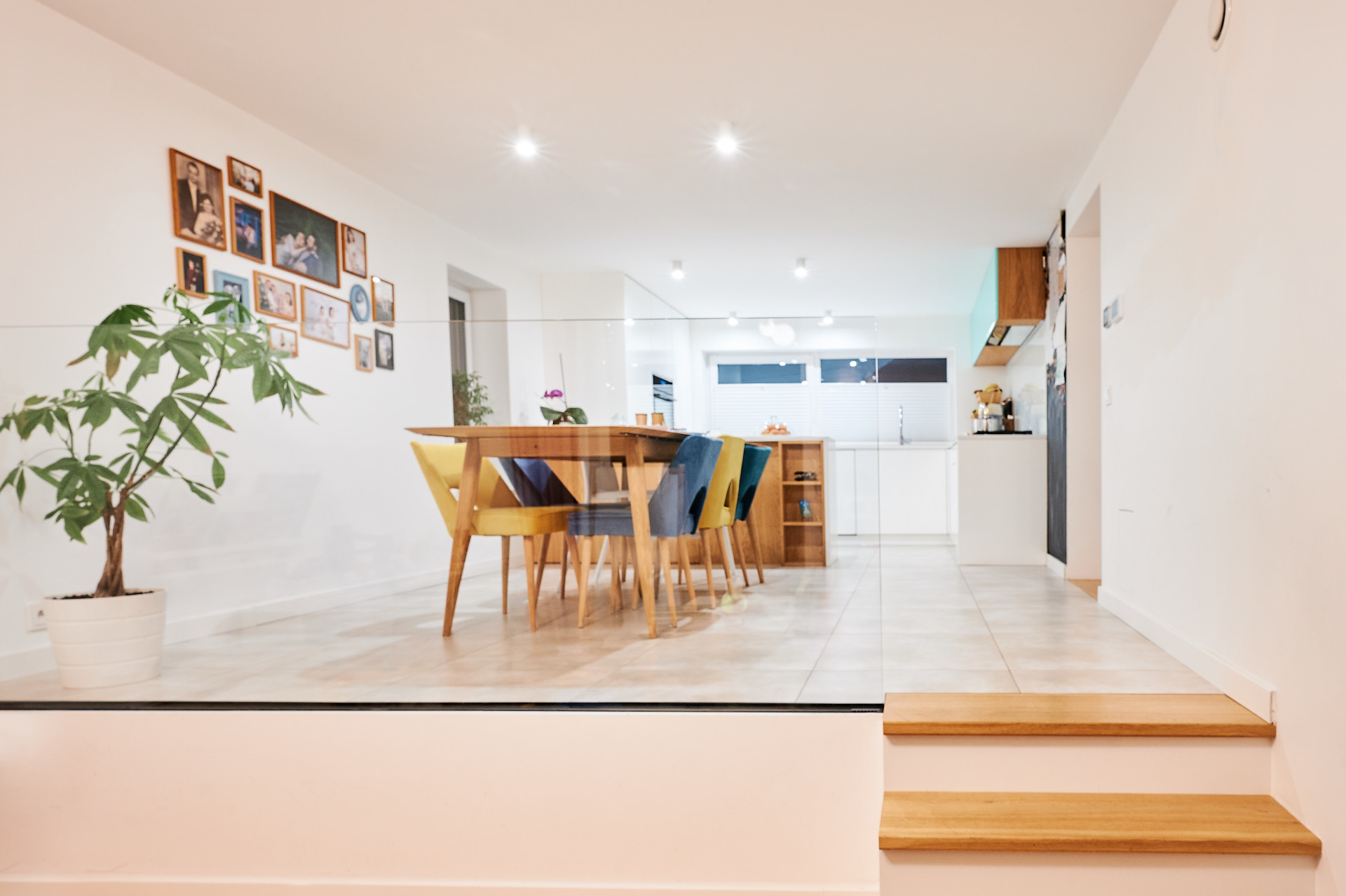 Nowoczesna jadalnia z drewnianym stołem, kolorowymi krzesłami, zdjęciami na ścianie i jasną kuchnią.